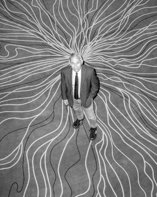 A person stands on a patterned carpet in a black and white photograph, shot from above with the person looking up at the camera.