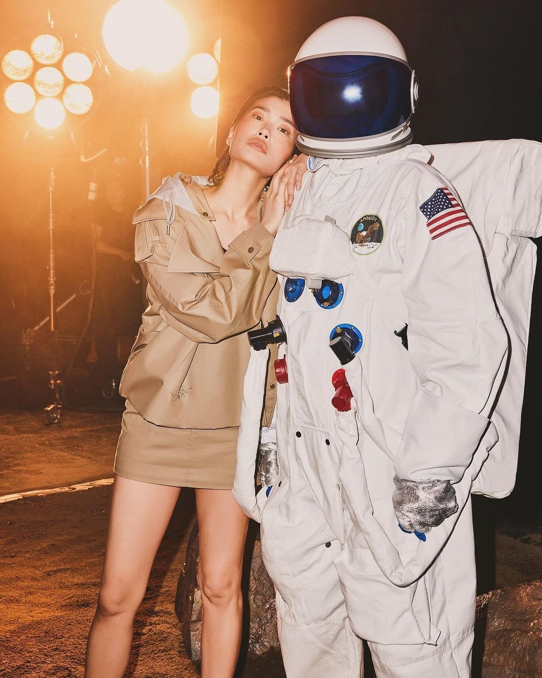 Woman poses next to an astronaut suit in a studio setting with soft lighting. She wears a stylish tan outfit with a matching skirt.