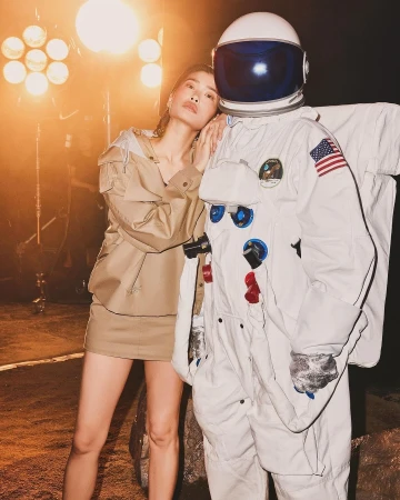 Woman poses next to an astronaut suit in a studio setting with soft lighting. She wears a stylish tan outfit with a matching skirt.