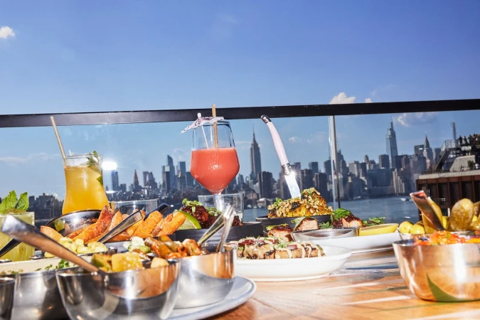 Drinks and appetizers are displayed on a rooftop with the New York City skyline in the background on a sunny day.
