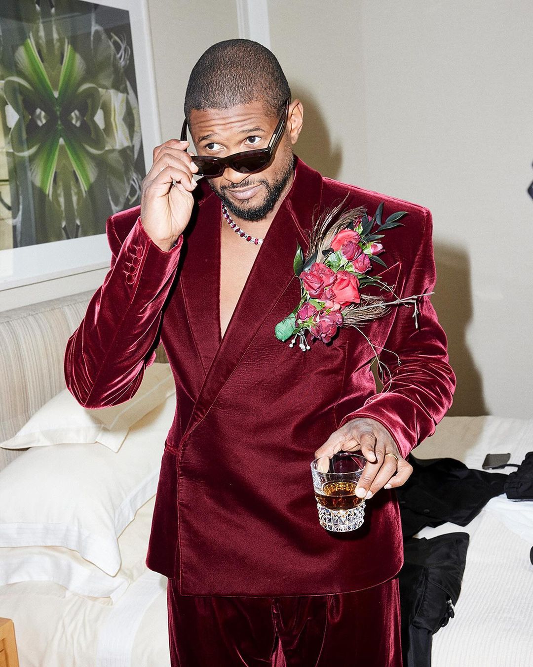 Adult male celebrity poses indoors wearing a maroon velvet suit, sunglasses, and a floral boutonniere, holding a glass with brown liquid.