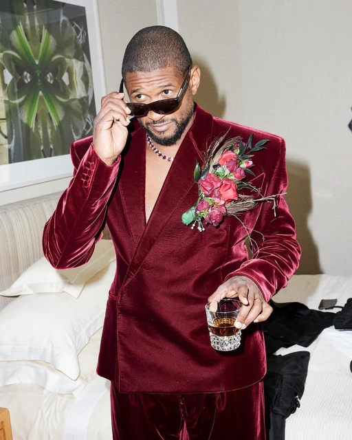 Adult male celebrity poses indoors wearing a maroon velvet suit, sunglasses, and a floral boutonniere, holding a glass with brown liquid.
