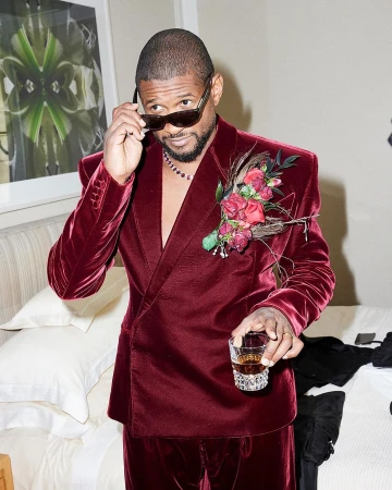 Adult male celebrity poses indoors wearing a maroon velvet suit, sunglasses, and a floral boutonniere, holding a glass with brown liquid.