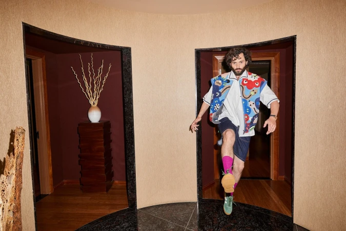 Man wearing a colourful patterned vest and shorts stepping through a doorway in a hallway by Jeremy Liebman.