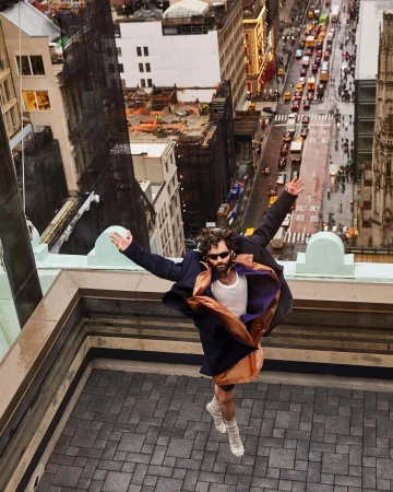 Man in motion on rooftop, arms outstretched, wearing an oversized coat and sunglasses with an urban city landscape in the background.