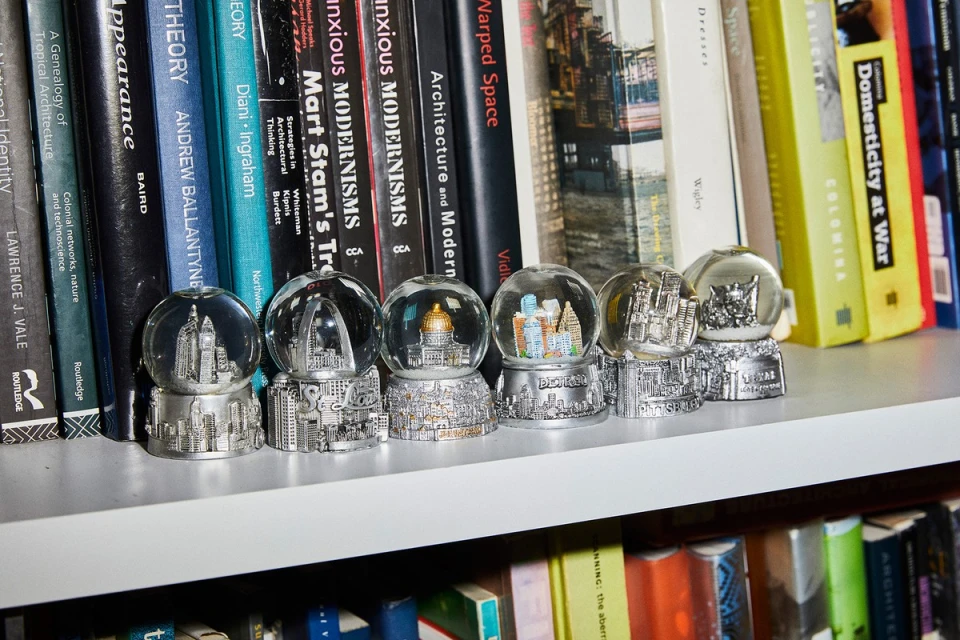 Seven snow globes with miniature cityscapes sit on a shelf amidst a backdrop of books in varying colors, arranged indoors.
