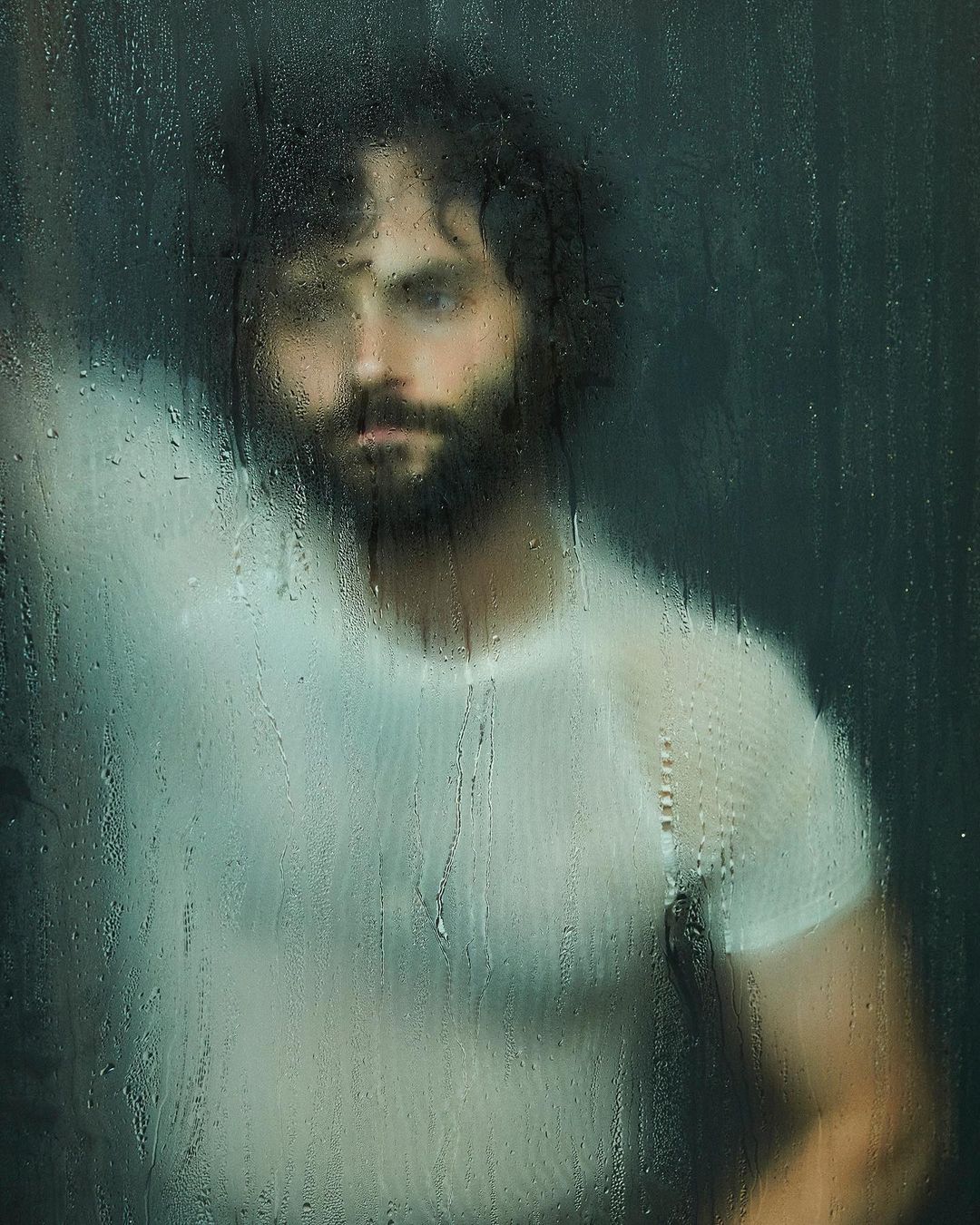 Portrait of a dark-haired adult male wearing a white t-shirt behind a wet glass pane with water droplets, creating a blurred effect.