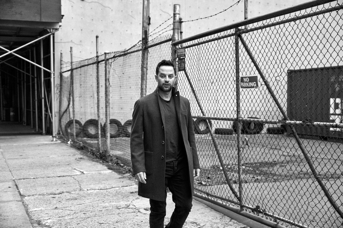 Black and white portrait of a man in a coat walking on a city street next to a fence and old building, offering a candid street-style.