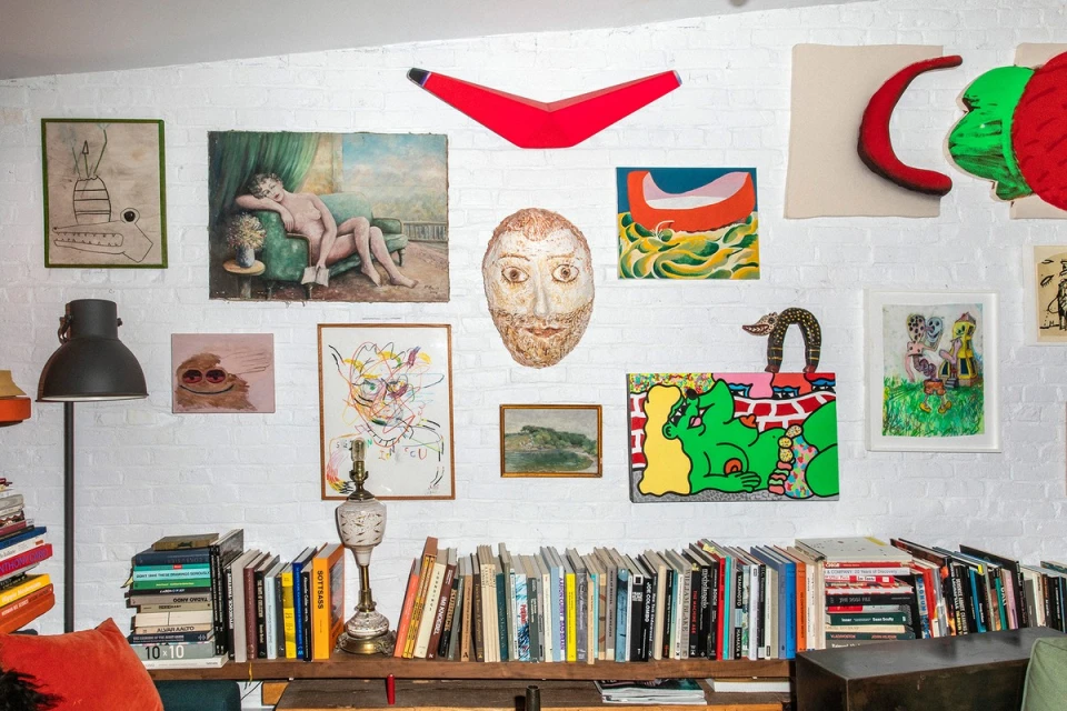 Living room interior with white brick wall decorated with colorful paintings and artwork above a shelf of books and decorative objects.