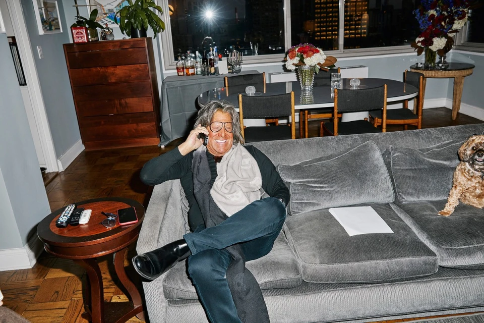 Adult male sitting on a grey couch indoors while talking on a cell phone, with a dog sitting beside him on the couch.