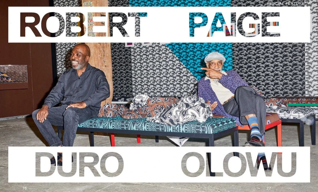 Two mature African American men are seated on a patterned bench in front of a wall of textiles. One man points to his right.
