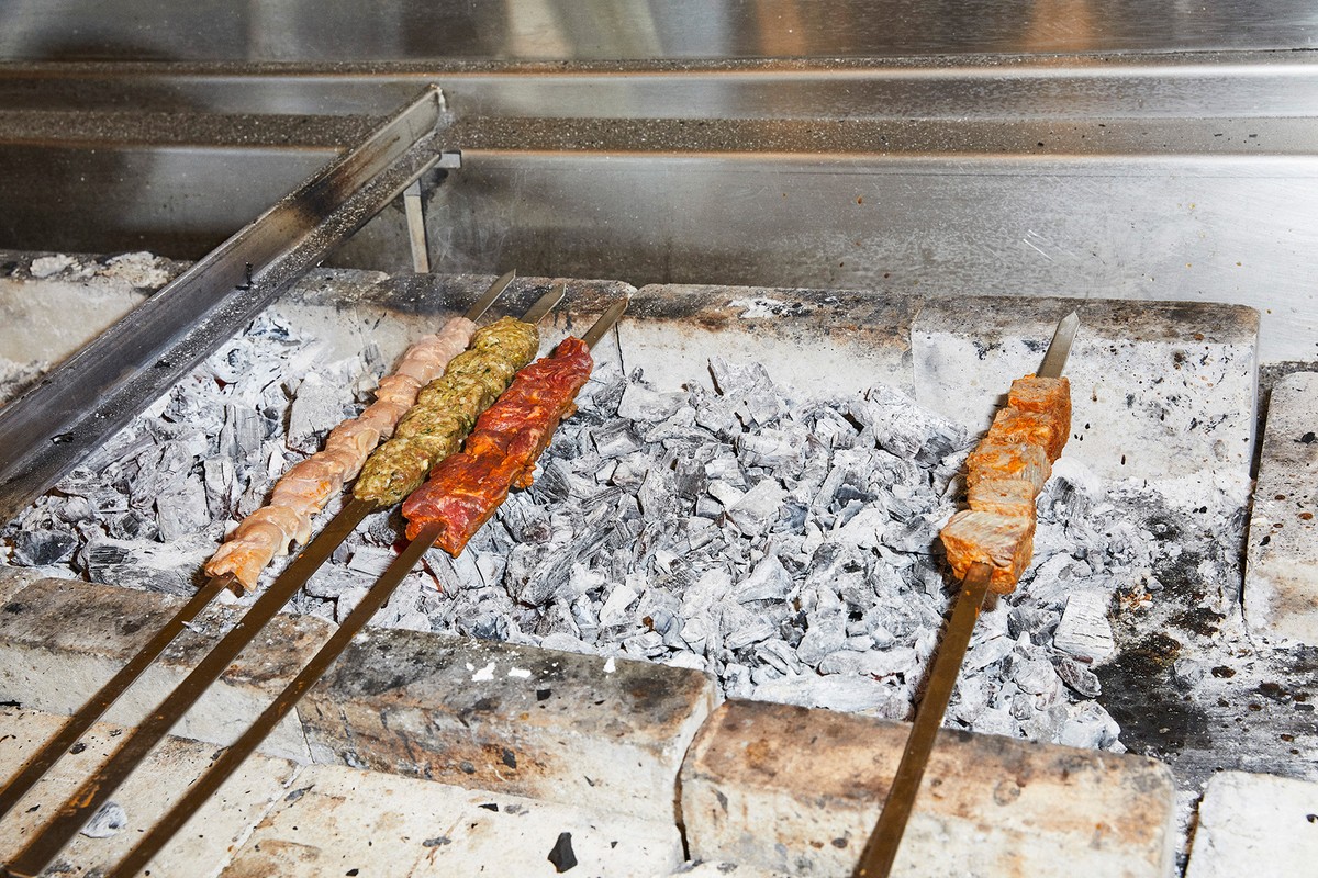 Skewers of seasoned meat are grilling over hot coals in a stone oven. The shish kebabs cook under a metal vent in an indoor setting.