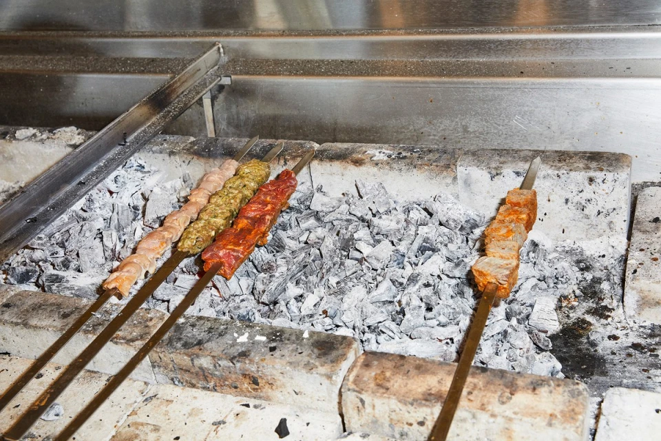 Skewers of seasoned meat are grilling over hot coals in a stone oven. The shish kebabs cook under a metal vent in an indoor setting.