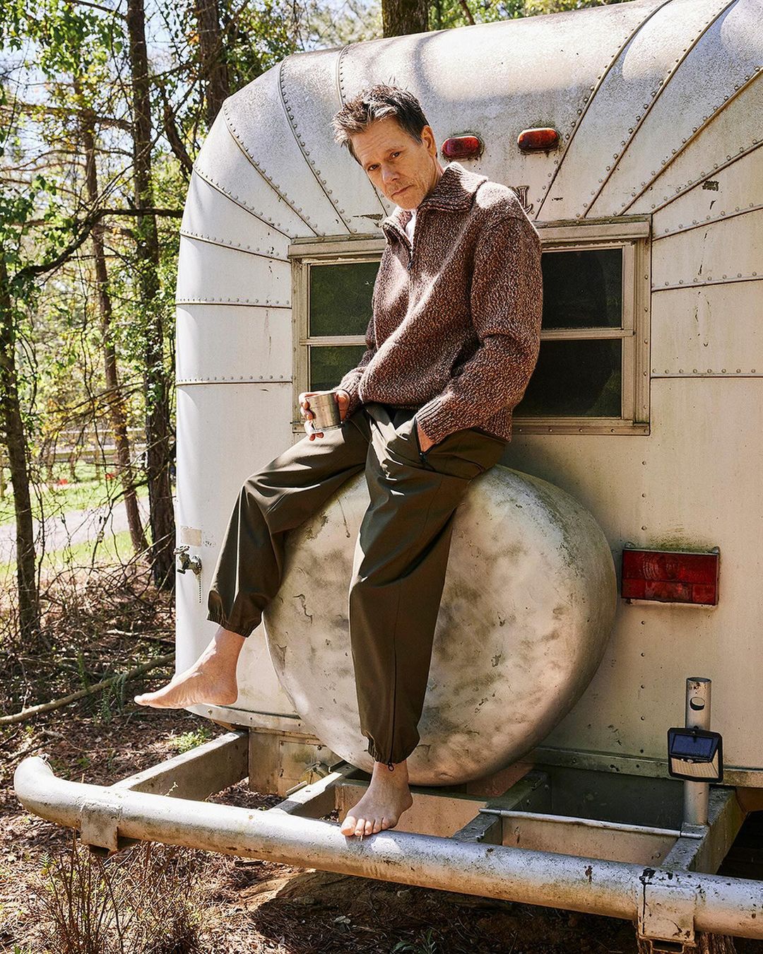 Adult male poses barefoot on a vintage travel trailer, holding a mug outside among trees. He wears olive pants and a wool sweater.