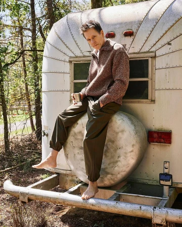 Adult male poses barefoot on a vintage travel trailer, holding a mug outside among trees. He wears olive pants and a wool sweater.