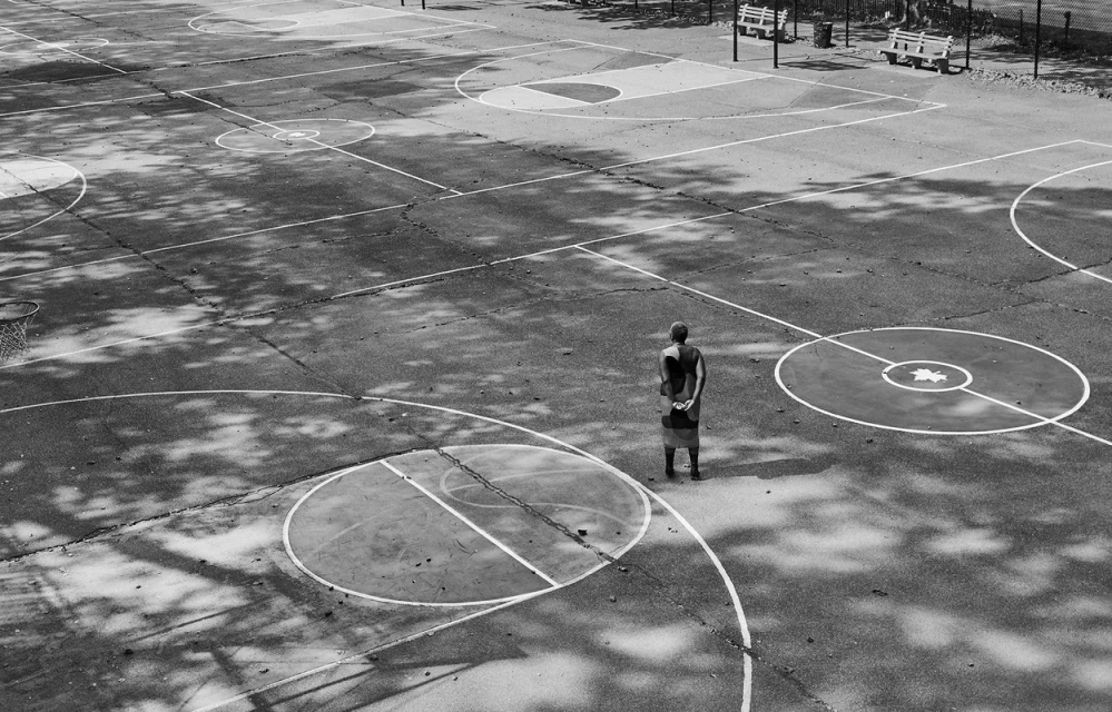 Monochrome image of an adult standing on an outdoor basketball court, surrounded by trees casting shadows, under daylight.
