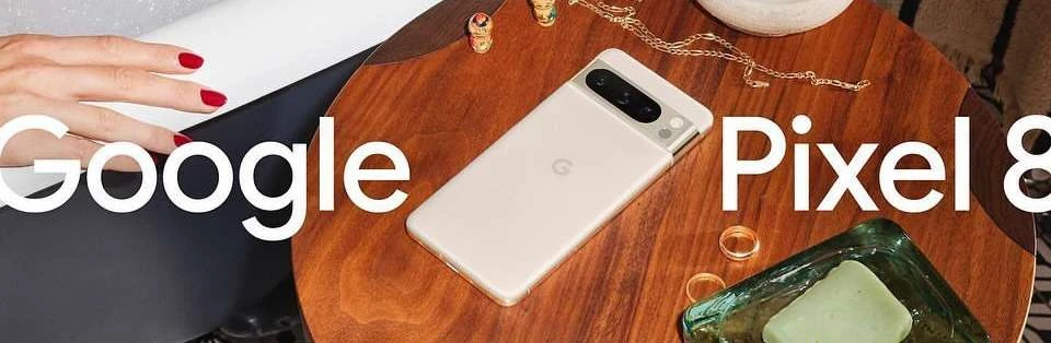 Google Pixel 8 smartphone product shot displayed on a wooden table with jewelry, next to a woman's hand with red nail polish.