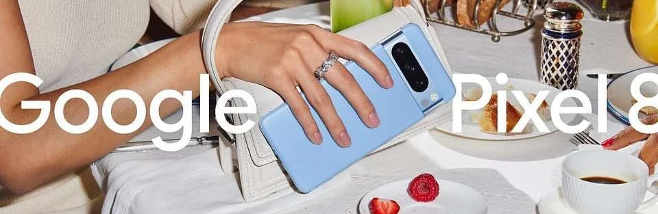 Product shot of a Google Pixel 8 smartphone in blue case, held by a hand with a ring, with breakfast and accessories on a table.