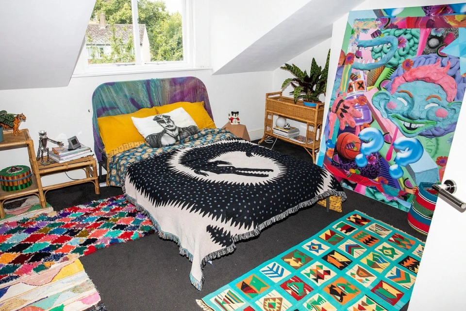 Interior bedroom view of a cozy bed with colorful throws, decorative pillows, and vibrant rugs, alongside eclectic wall art and plants.