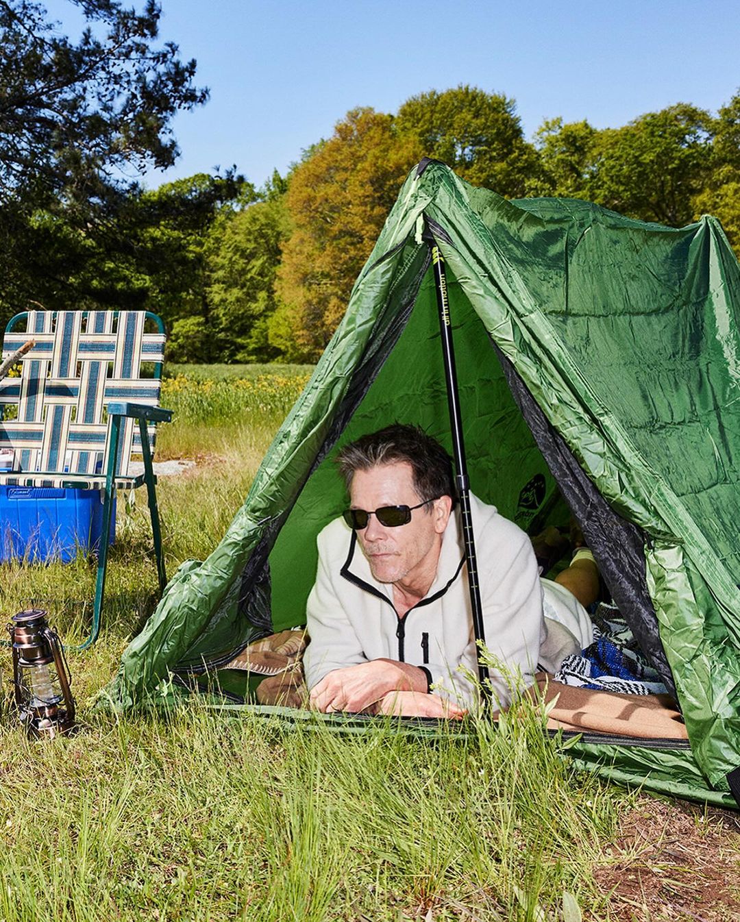 Man in a green tent with a camping lantern, folding chair, blue bucket, and trees in the background on a clear sunny day.