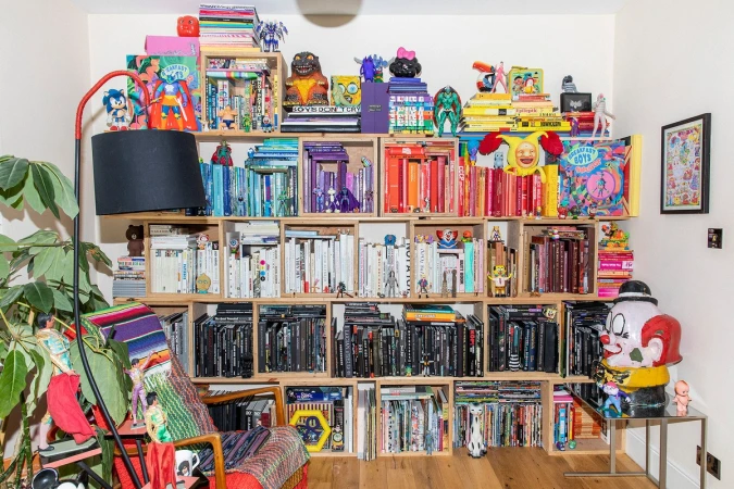 Colorful book collection artfully arranged on a shelving unit, complete with figures and decorative objects, accented by a floor lamp and plant.