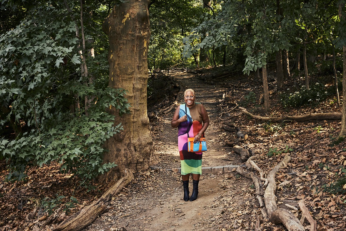 Adult woman poses on a wooded path wearing a colorful dress and carrying a handbag. Trees and leafy foliage in the background.