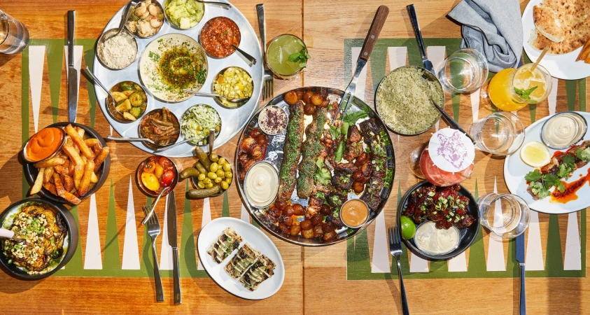 Overhead shot of a table laden with various Middle Eastern dishes, including hummus, salads, grilled meats, fries, and colorful cocktails.