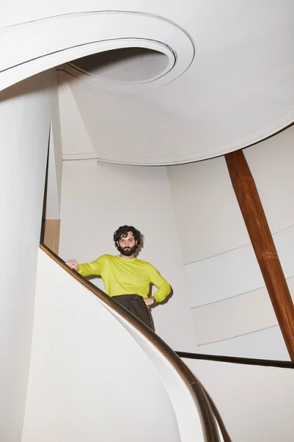 Person in chartreuse sweater and dark pants poses on a modern white spiral staircase with wooden accents in a bright indoor setting.