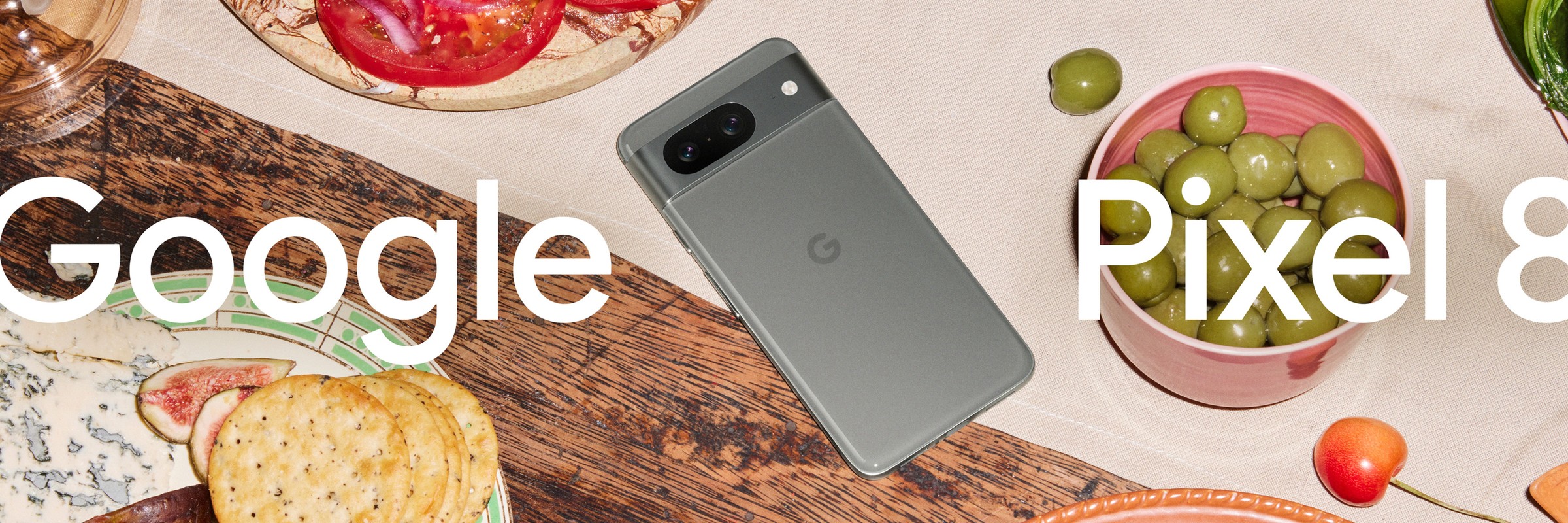 Google Pixel 8 smartphone with food arrangement: olives, tomatoes, figs, and crackers on a wooden and linen surface.