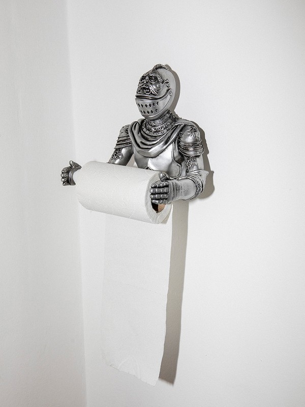 A silver knight figure holds a toilet paper roll in a brightly lit bathroom setting, creating a whimsical and unexpected contrast.