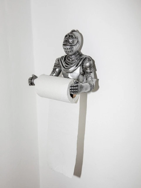 A silver knight figure holds a toilet paper roll in a brightly lit bathroom setting, creating a whimsical and unexpected contrast.