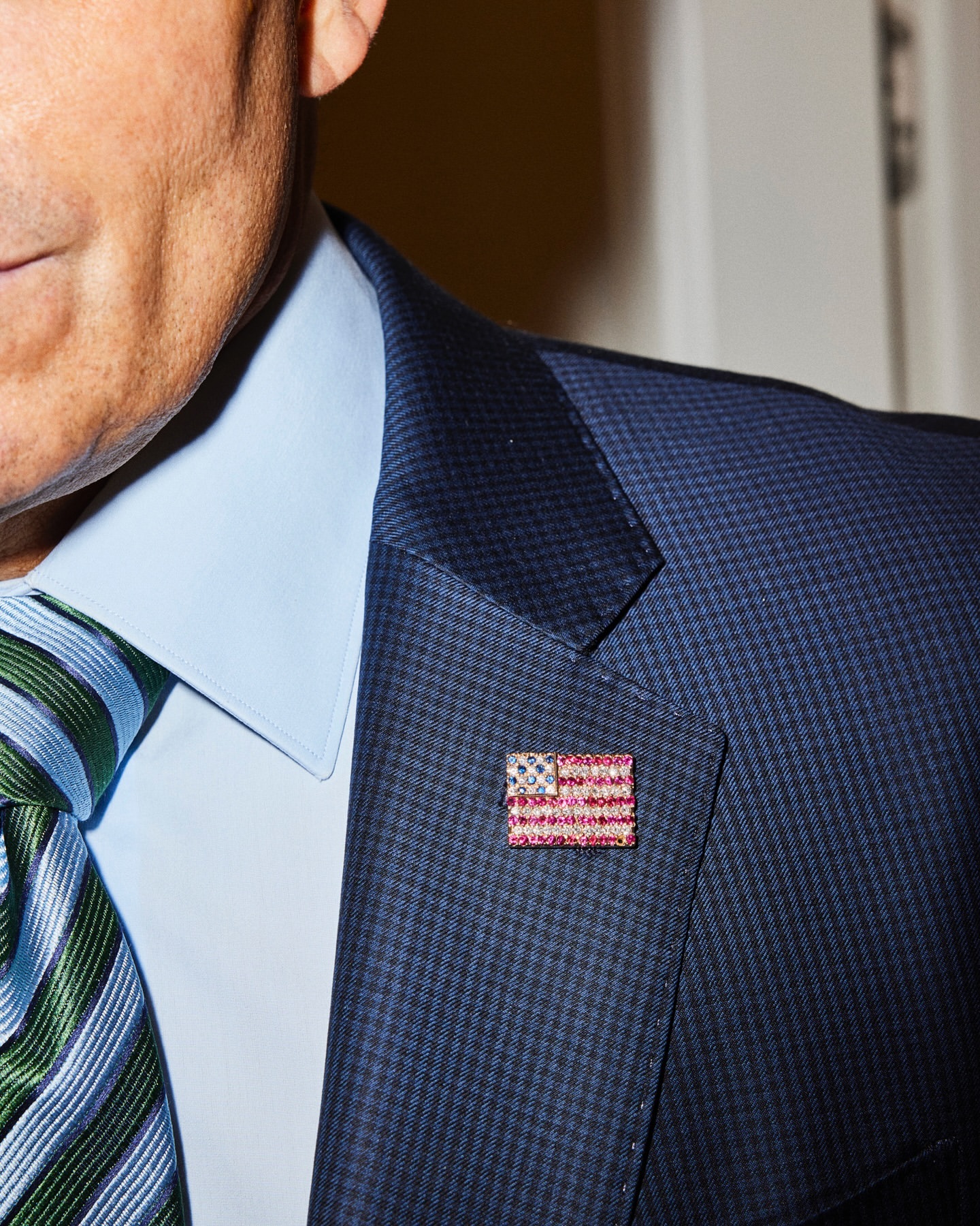 Close-up of a man wearing a suit with an American flag pin on the lapel, a light blue shirt, and a green and blue striped tie.