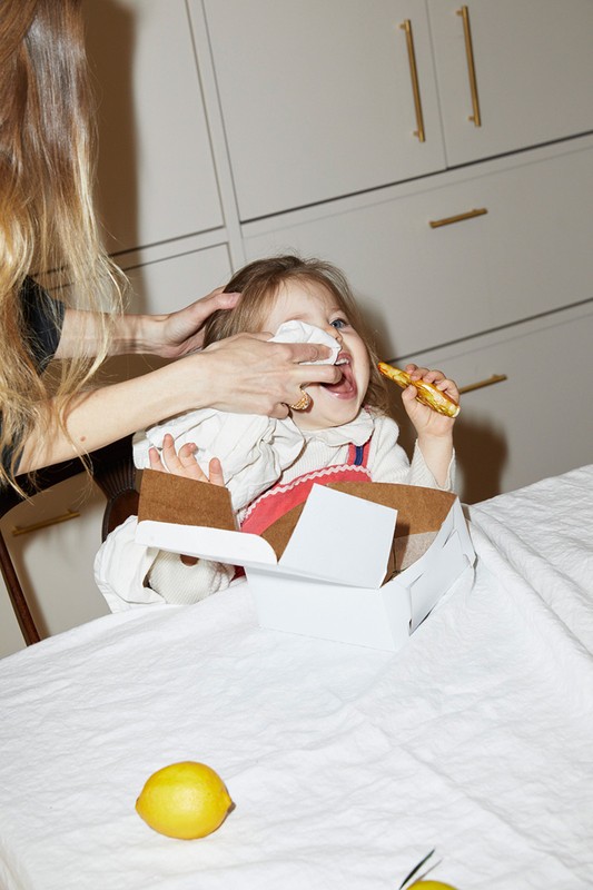 Indoor shot captures a child eating with help from an adult, sitting at a table with a white tablecloth, food box, and lemons.