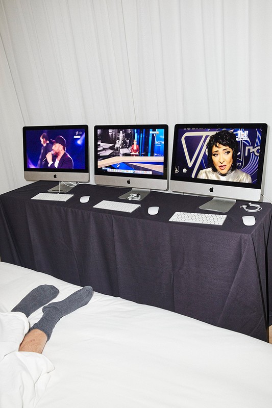 Three computer monitors showing different presenters on top of a table with a black tablecloth. Person's legs and feet on a white bedsheet in foreground.