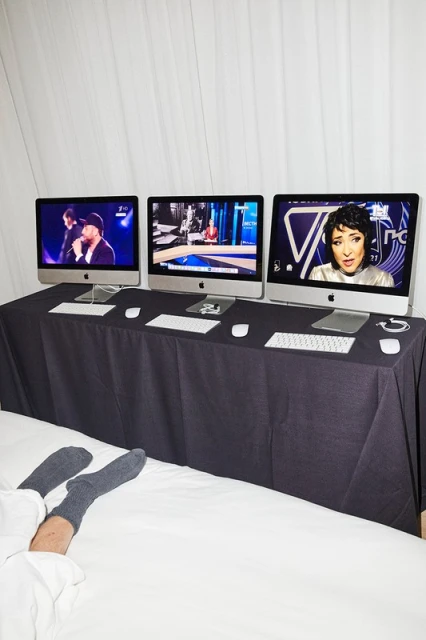 Three computer monitors showing different presenters on top of a table with a black tablecloth. Person's legs and feet on a white bedsheet in foreground.