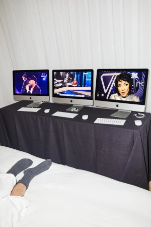 Three computer monitors showing different presenters on top of a table with a black tablecloth. Person's legs and feet on a white bedsheet in foreground.