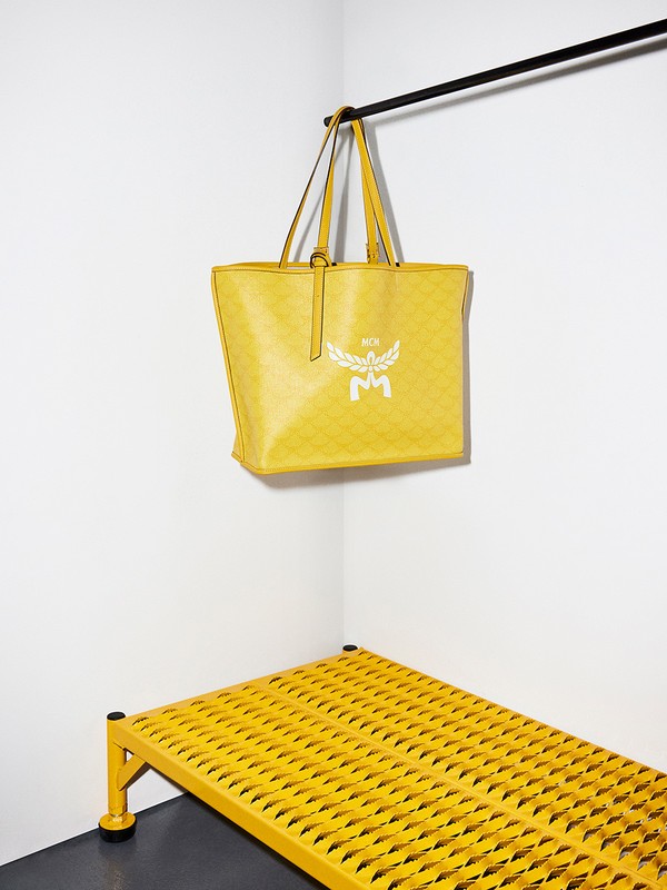 Studio shot of a yellow MCM tote bag hanging from a black bar, with a matching yellow metal grid shelf in the corner against white walls.