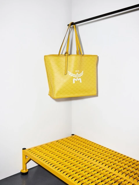 Studio shot of a yellow MCM tote bag hanging from a black bar, with a matching yellow metal grid shelf in the corner against white walls.