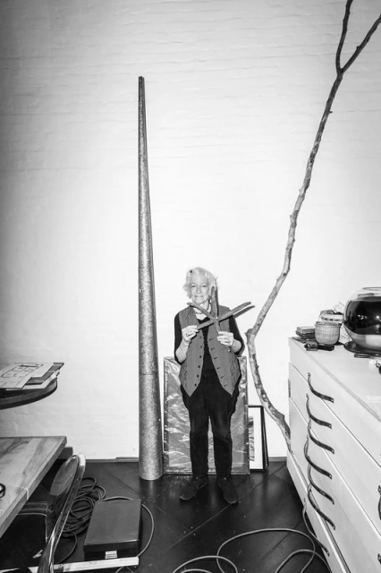 Black-and-white shot of an adult woman standing indoors holding an object, with art and furniture visible. Full shot, eye-level.