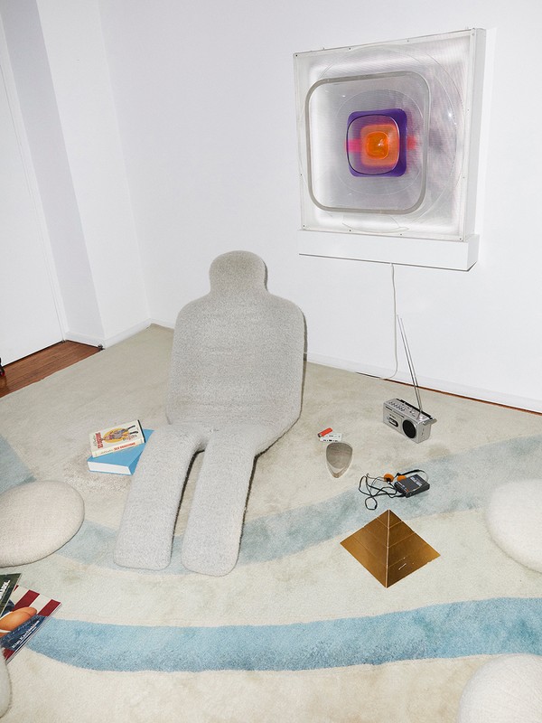 Unique interior shot featuring a gray, human-shaped chair on a patterned rug, accompanied by books, a retro radio, and a modern wall light.