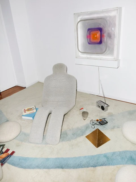 Unique interior shot featuring a gray, human-shaped chair on a patterned rug, accompanied by books, a retro radio, and a modern wall light.