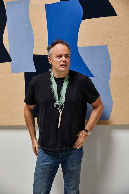 Adult male poses in front of a blue and beige abstract art piece with scissors around his neck, wearing a black t-shirt and jeans.