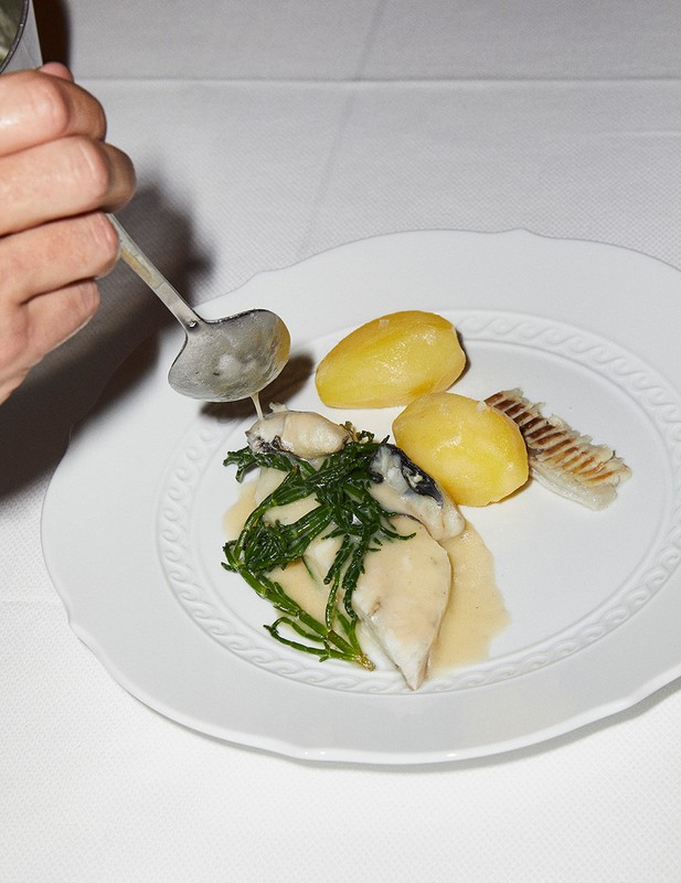 Overhead shot of a plate with fish, potatoes, and greens, drizzled with sauce from a ladle, highlighting food presentation and culinary preparation.