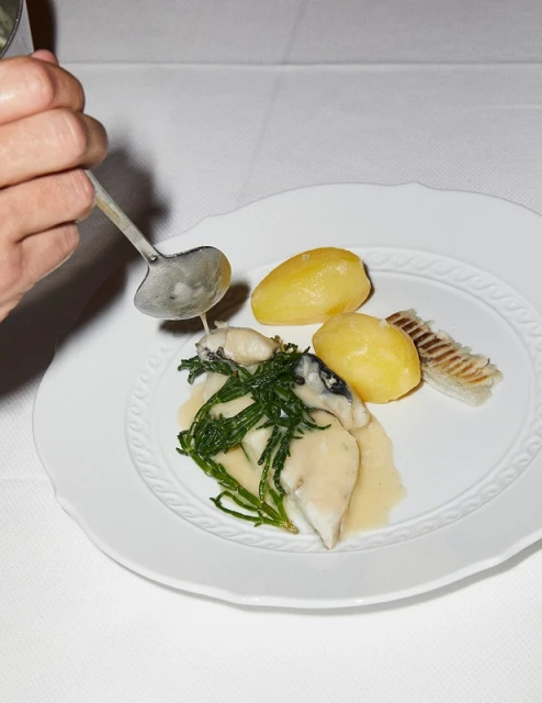 Overhead shot of a plate with fish, potatoes, and greens, drizzled with sauce from a ladle, highlighting food presentation and culinary preparation.