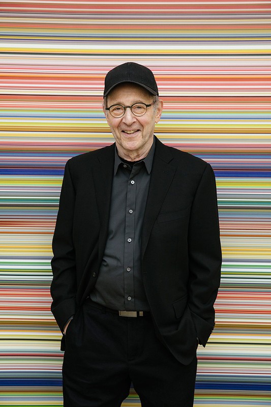An elderly man with glasses and a baseball cap in a black suit stands in front of a colorful horizontal striped background.