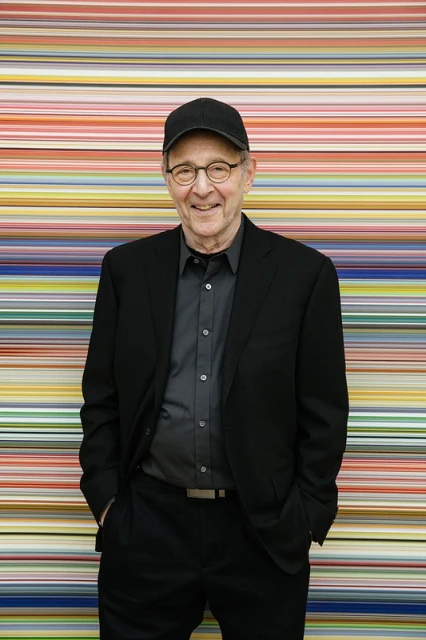 An elderly man with glasses and a baseball cap in a black suit stands in front of a colorful horizontal striped background.