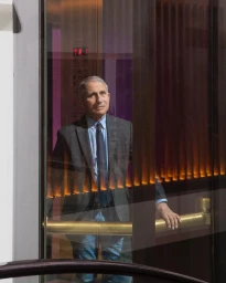 Anthony Fauci standing inside a glass elevator wearing a charcoal suit jacket and blue tie, photographed by Jeremy Liebman.