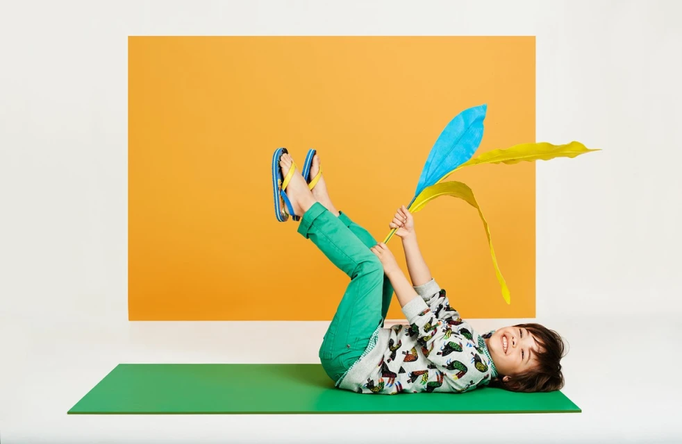 Child with green pants and colorful top lying on green mat, holding up blue and yellow artificial leaves in front of orange backdrop.