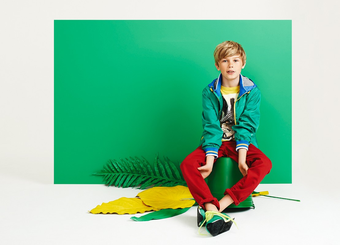 Blond boy in a green jacket sits on a green bucket in a studio with green and yellow painted leaves. Red pants and colorful sneakers.