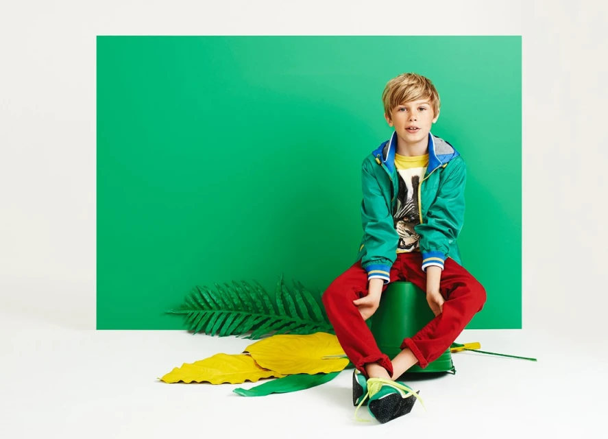 Blond boy in a green jacket sits on a green bucket in a studio with green and yellow painted leaves. Red pants and colorful sneakers.